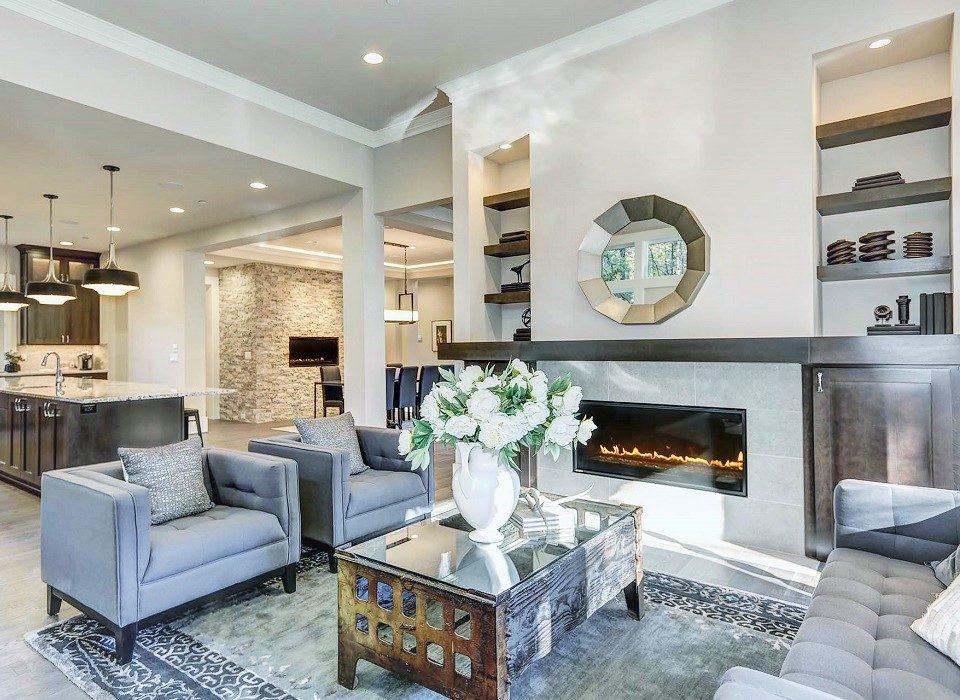 Chic living room filled with built-in cabinets flanking round mirror atop grey tile fireplace, tufted sofa facing two armchairs and window wall overlooking  lush outdoors. Northwest, USA